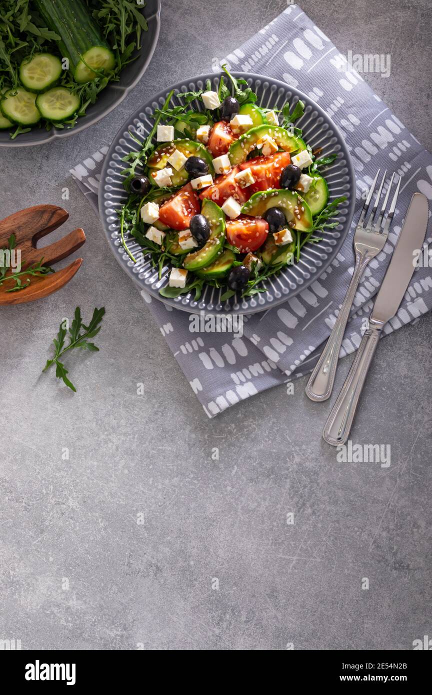 Fresh vegetarian salad of rocket, cucumber, tomatoes, cheese, avocado and olives garnished with balsamic glaze and sesame seeds on grey stone Stock Photo