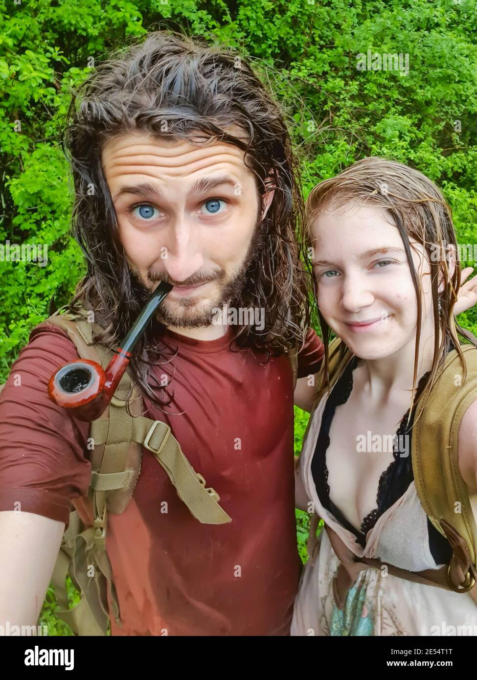 Selfie shot of a young couple drenched wet from a sudden rain in the middle of a hike in the forest, smiling at camera and the guy smoking a pipe. Stock Photo
