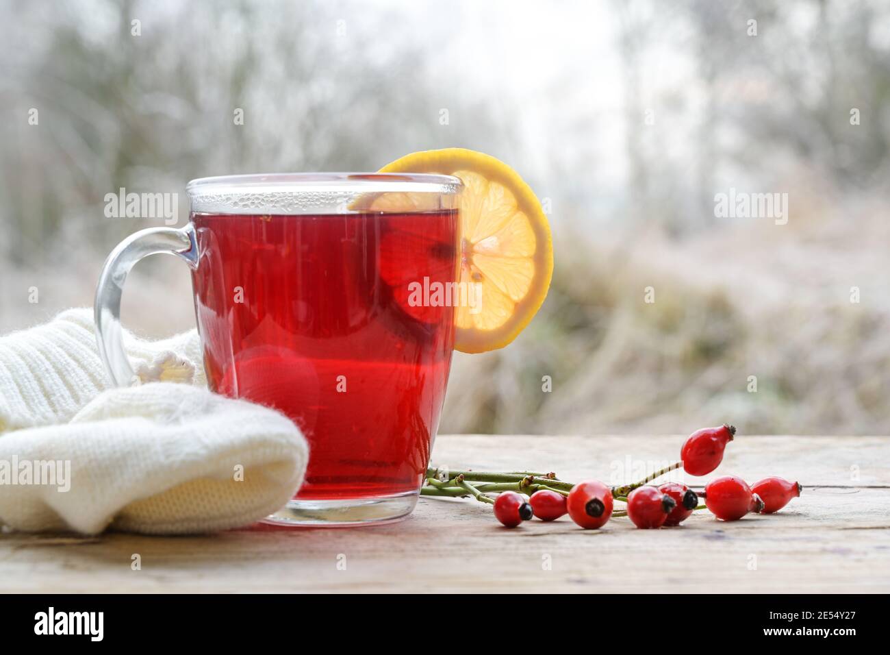 Hot red rose hip tea with a lemon slice in a glass mug on a rustic wooden table against a frosty winter landscape, copy space, selected focus, narrow Stock Photo