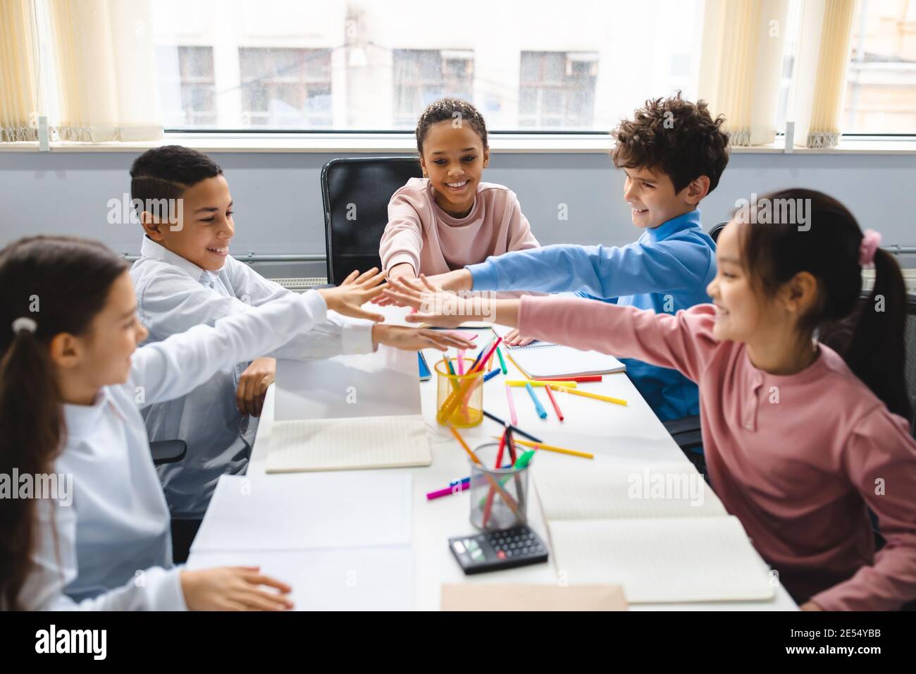 Diverse small schoolchildren putting hands together at classroom Stock Photo