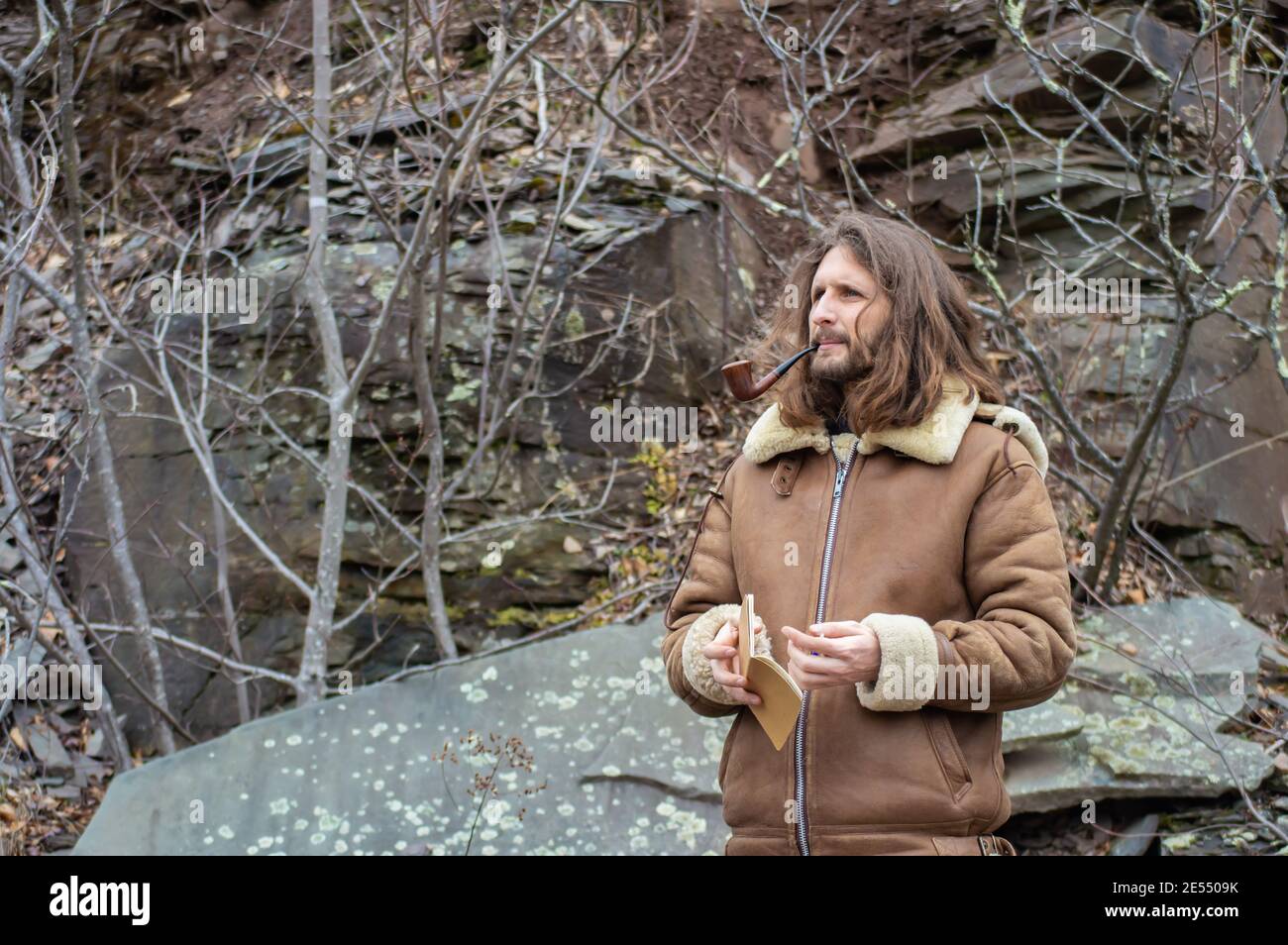 Young man with long hair walking in the woodlands with a sheepskin coat, a paper notebook and smoking a wooden vintage Churchwarden pipe. Stock Photo