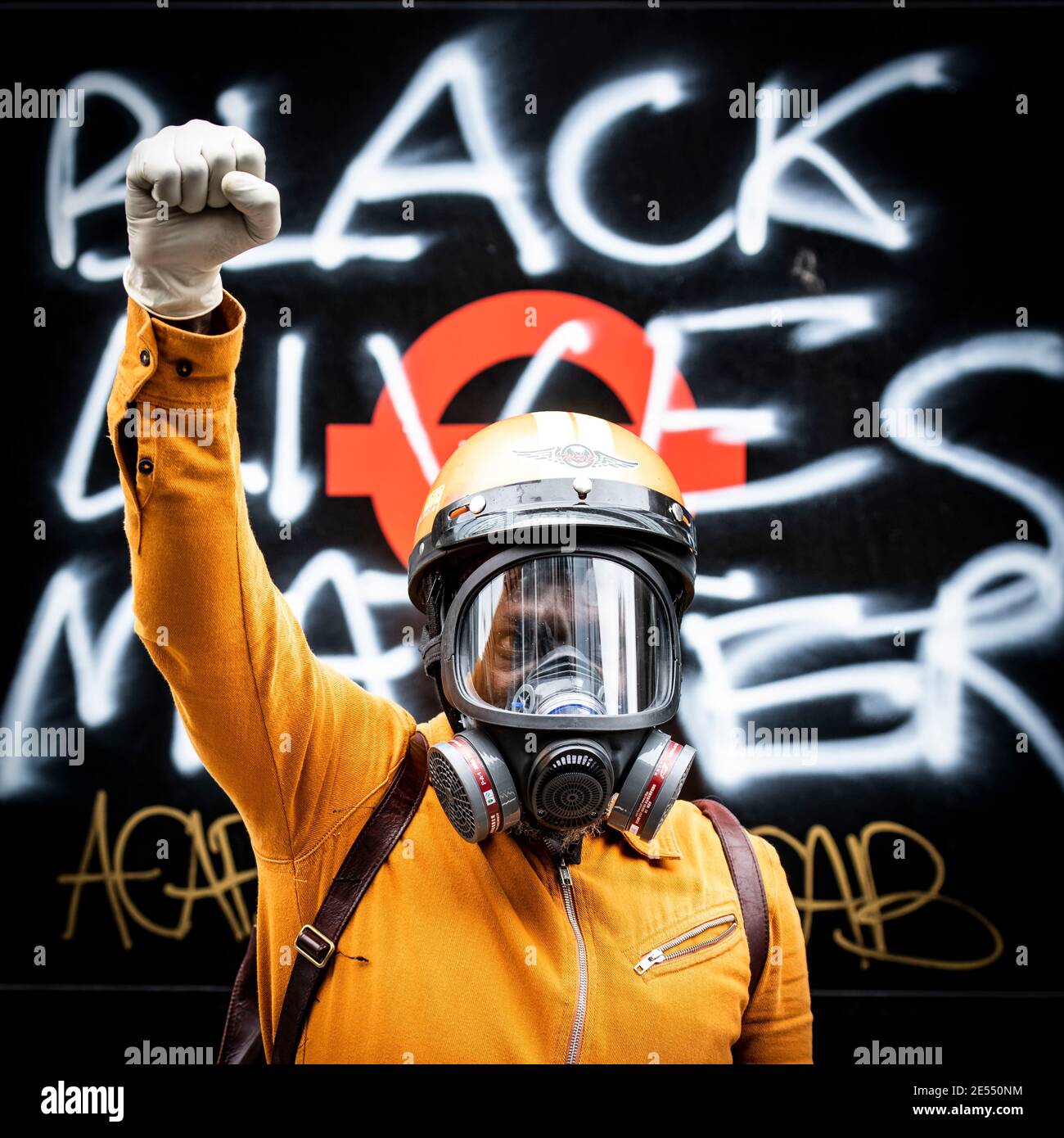 Black Lives Matter protestor wearing full ppe face mask raises a clenched fist in front of a bus shelter which has black lives matter sprayed on the side Stock Photo