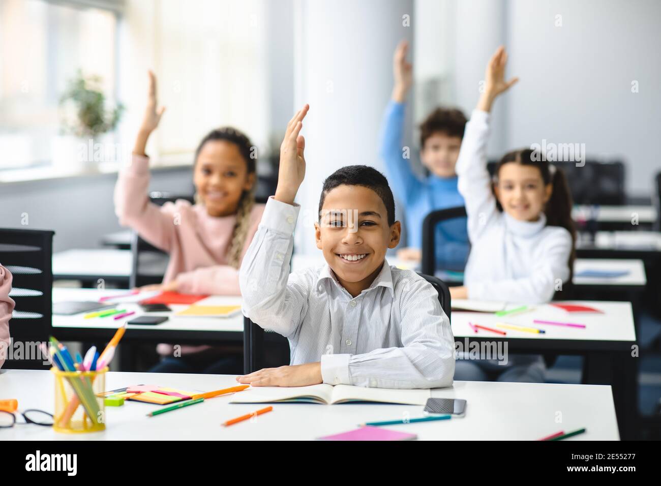 Diverse small schoolkids raising hands at classroom Stock Photo