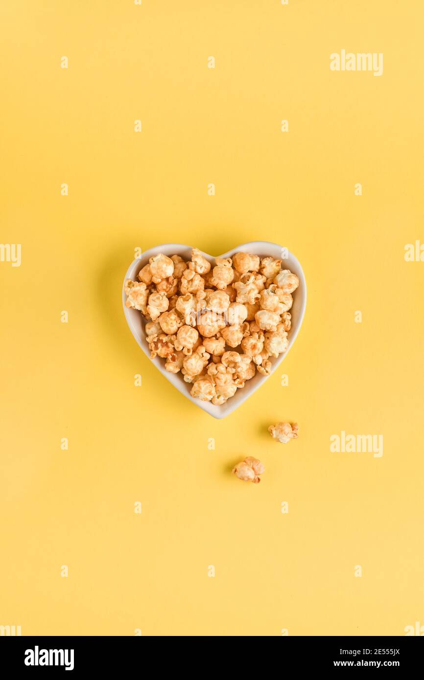 Delicious sweet popcorn with caramel in white ceramic heart plate, isolated on trend color yellow background. Stock Photo