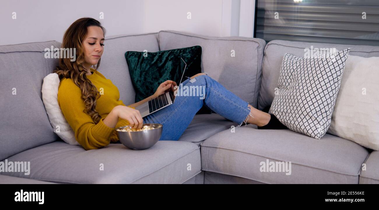 Young woman lies on the couch with her laptop and eats popcorn Stock ...