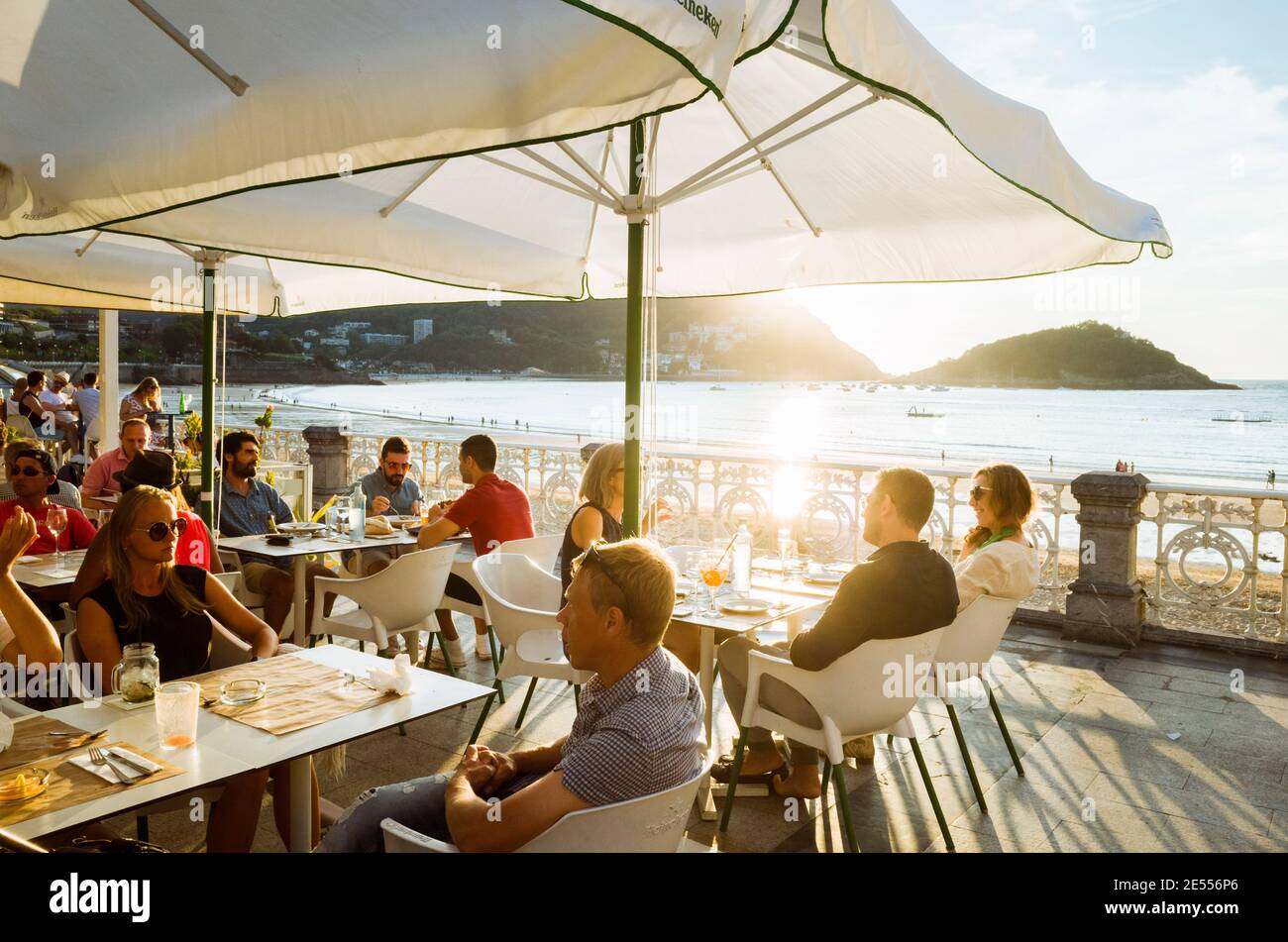 Donostia, Gipuzkoa, Basque Country, Spain - July 15th, 2019 : People relax at sunset in a chic sidewalk cafe in the promenade of La Concha beach. Stock Photo