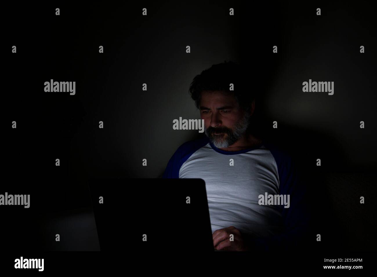 Bearded man sitting on sofa at home at night, working on laptop. Stock Photo