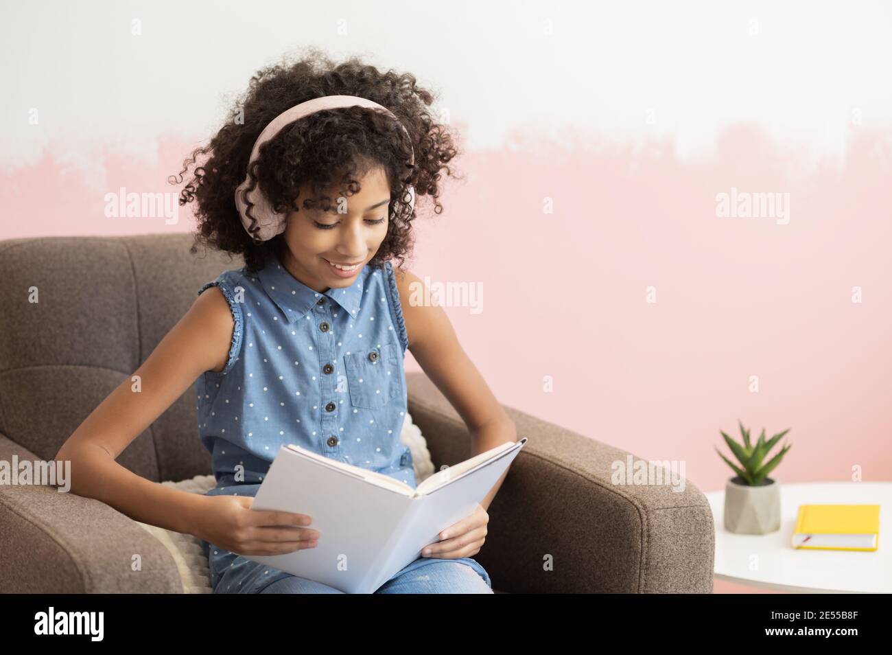 A clever African preteen school girl spending leisure time with a book reading sitting at armchair at home Stock Photo
