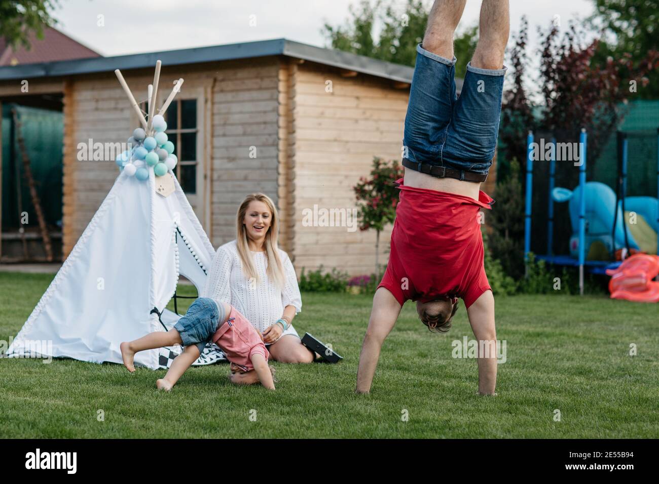 Happy family spending time together outside in the garden. Child having fun with his parents doing headstands on the lawn. Stock Photo