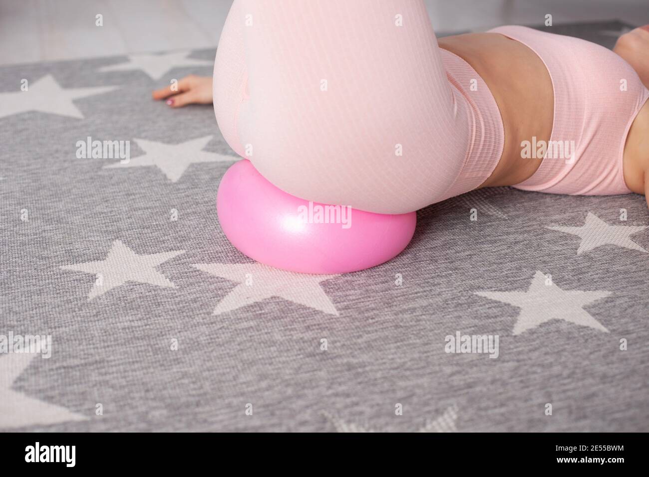 close-up woman sports booty in pink pants lying on a rubber flattened pilates ball on a gray carpet with white stars Stock Photo