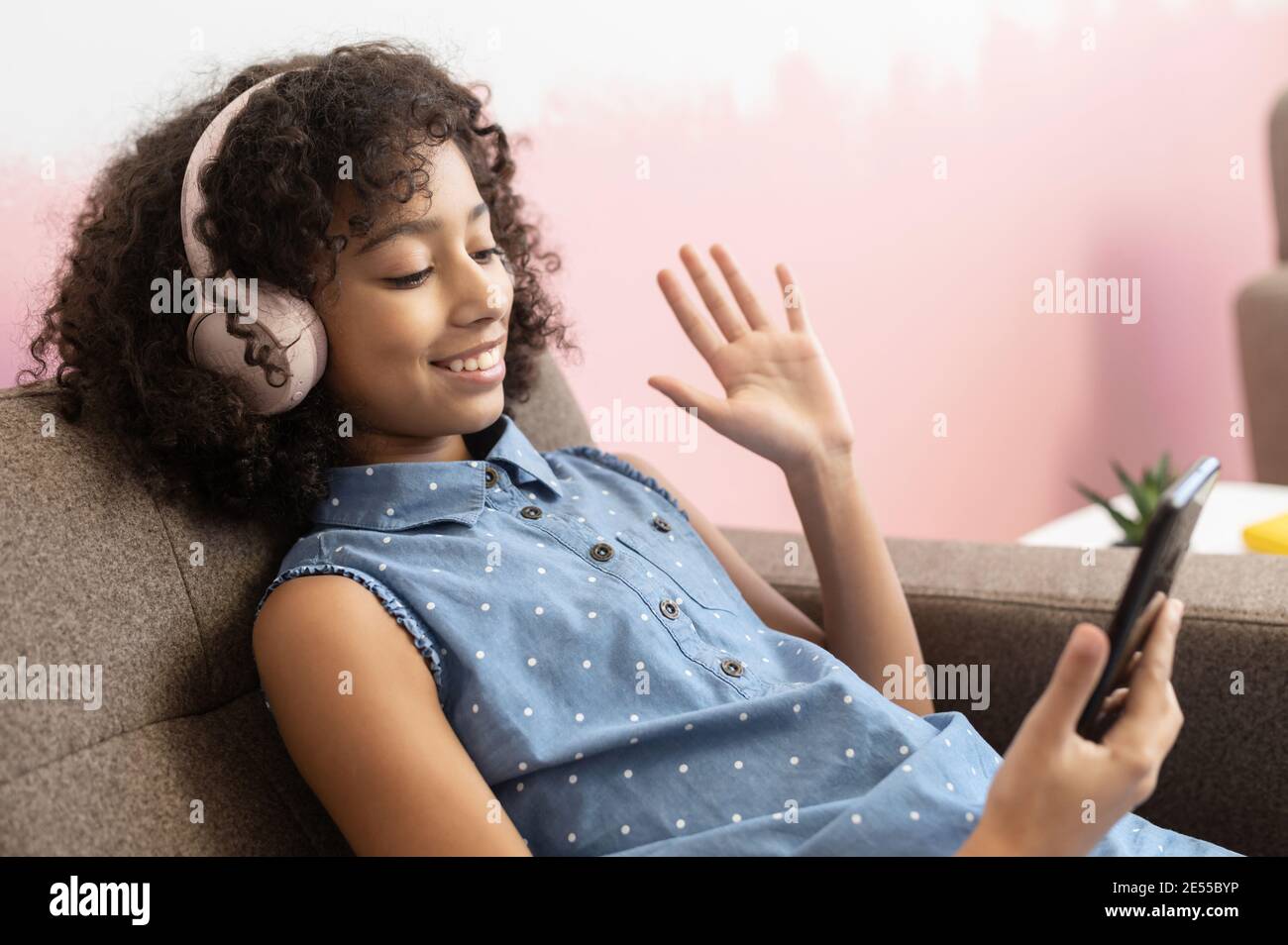 Pretty African preteen schoolgirl sitting at armchair with a smartphone at home, waving hi into webcam, chatting online with a friends, classmates Stock Photo
