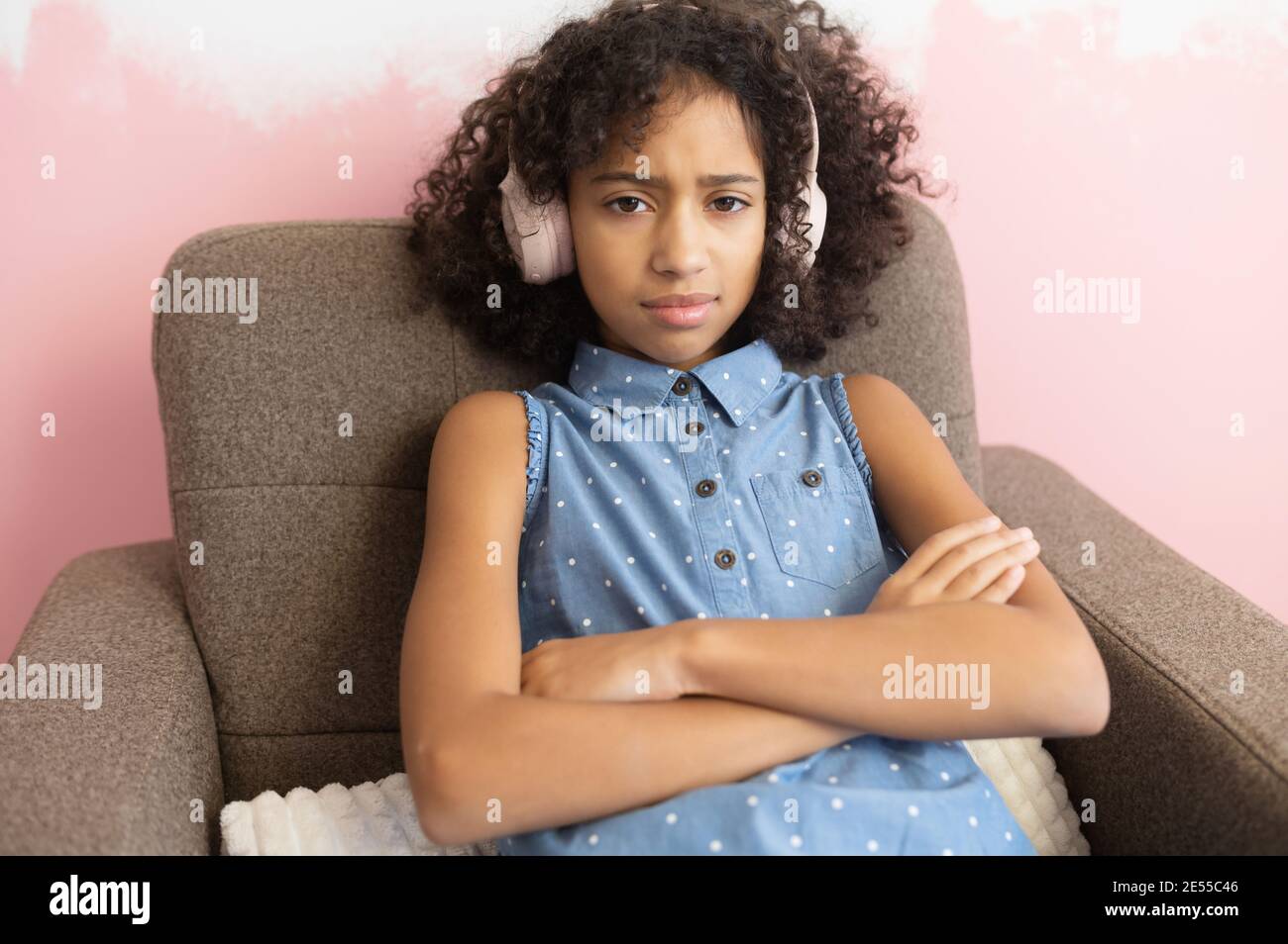 A curly African-American school-age girl wearing headphones sits on the armchair frowning face in displeasure, keeps arms folded. Difficult teen-age Stock Photo