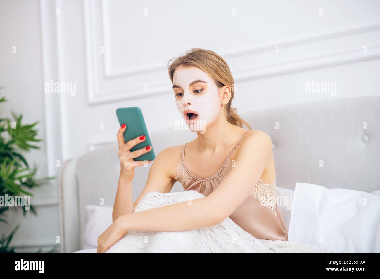 Blonde girl with a mask on her face sitting in bed with smartphone in hand Stock Photo