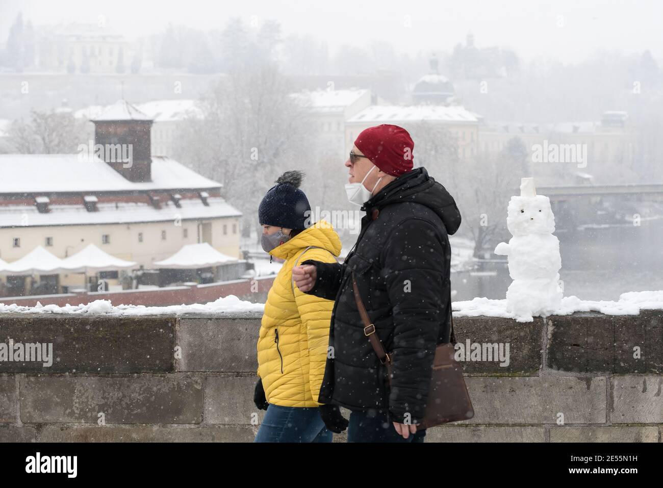 A couple wearing face masks as a preventive measure against the spread of coronavirus walk past a snowman on Charles Bridge during snowfall. Heavy snowfall is observed in the Prague, historical city center, which is part of the Unesco heritage, and iconic Charles bridge. Stock Photo
