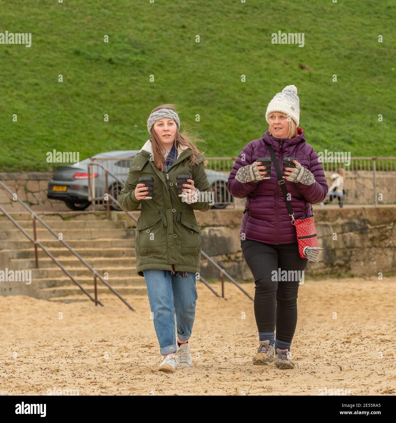 Young woman (20s) and older woman (50s) dressed for the cold carry four cups of coffee over sand beach towards the camera. Stock Photo