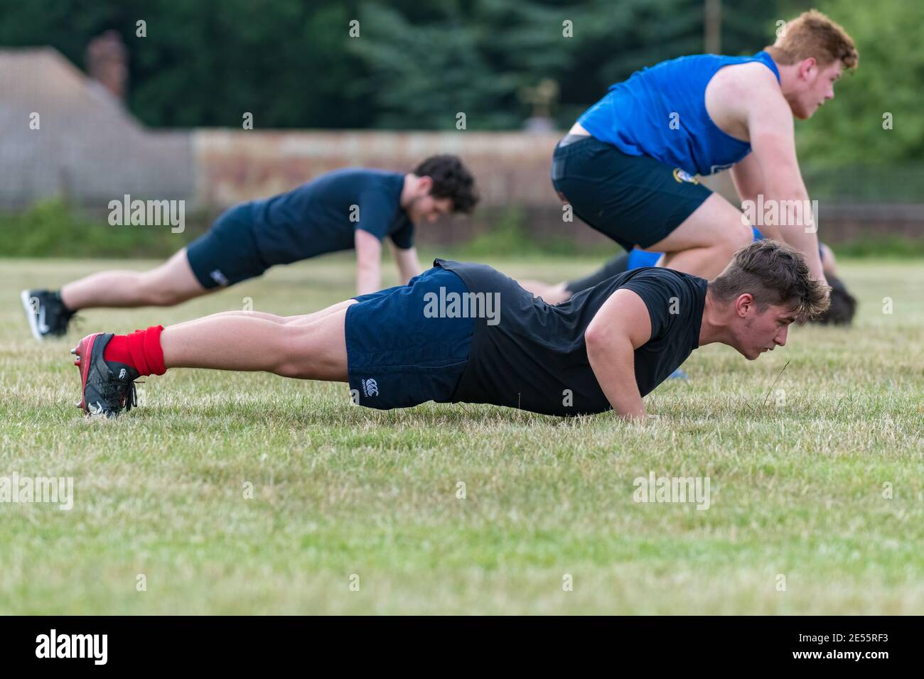 Three young men (late teens) wearing shorts and t-shirts doing press ups and burpees on grass field. Stock Photo