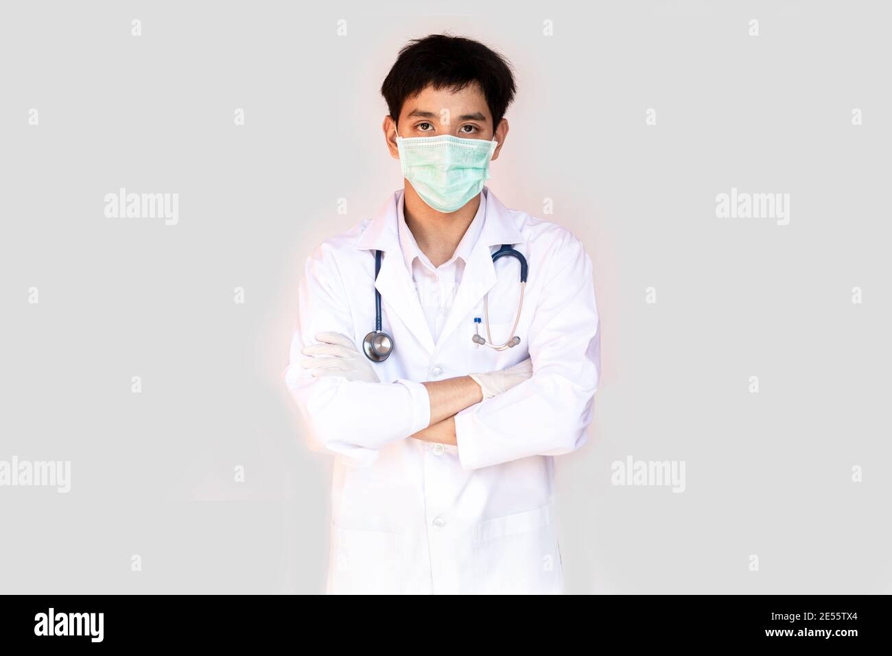 A doctor posing with arms crossed on a white background is wearing a medical face mask and stethoscope. Young Asian doctor wearing a white coat. Stock Photo