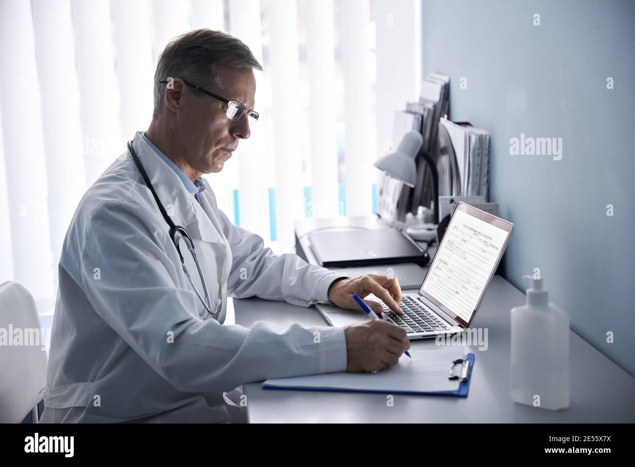 Serious mature doctor writing notes, using laptop in medical office. Stock Photo