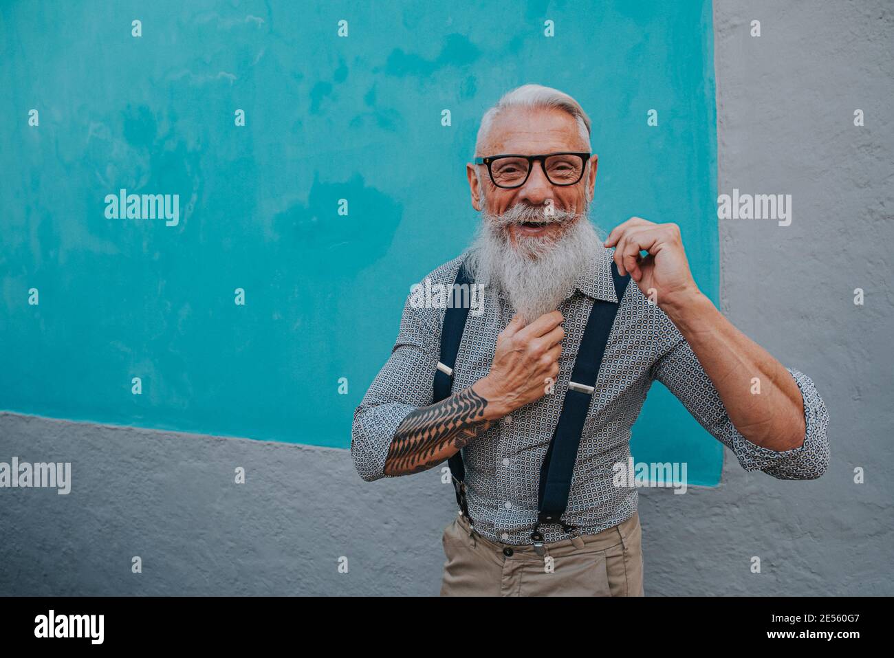an older man in hipster clothes and glasses and a long white beard poses on a blue wall Stock Photo