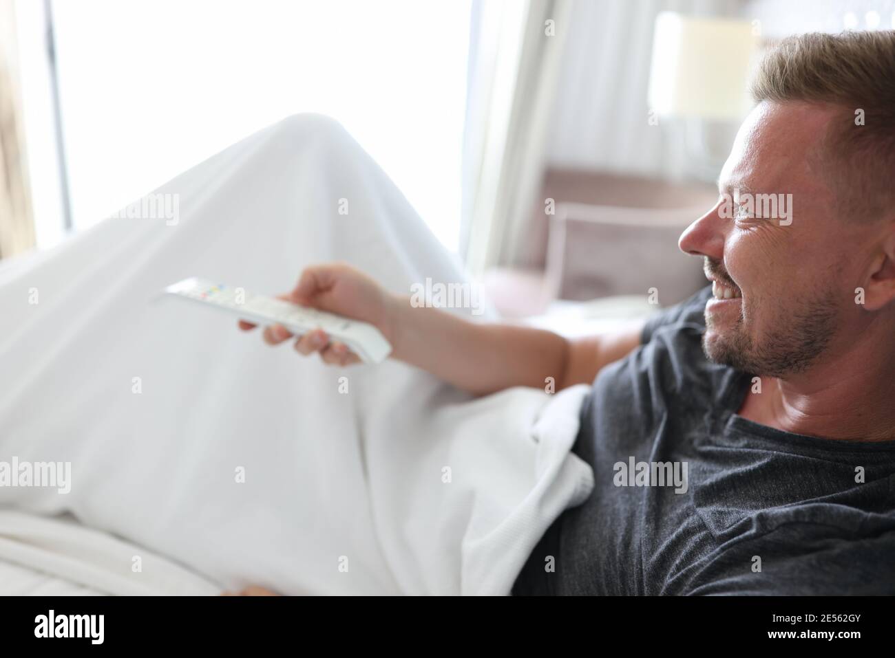 Smiling Man Lies In Bed And Presses Buttons On Remote Control Stock