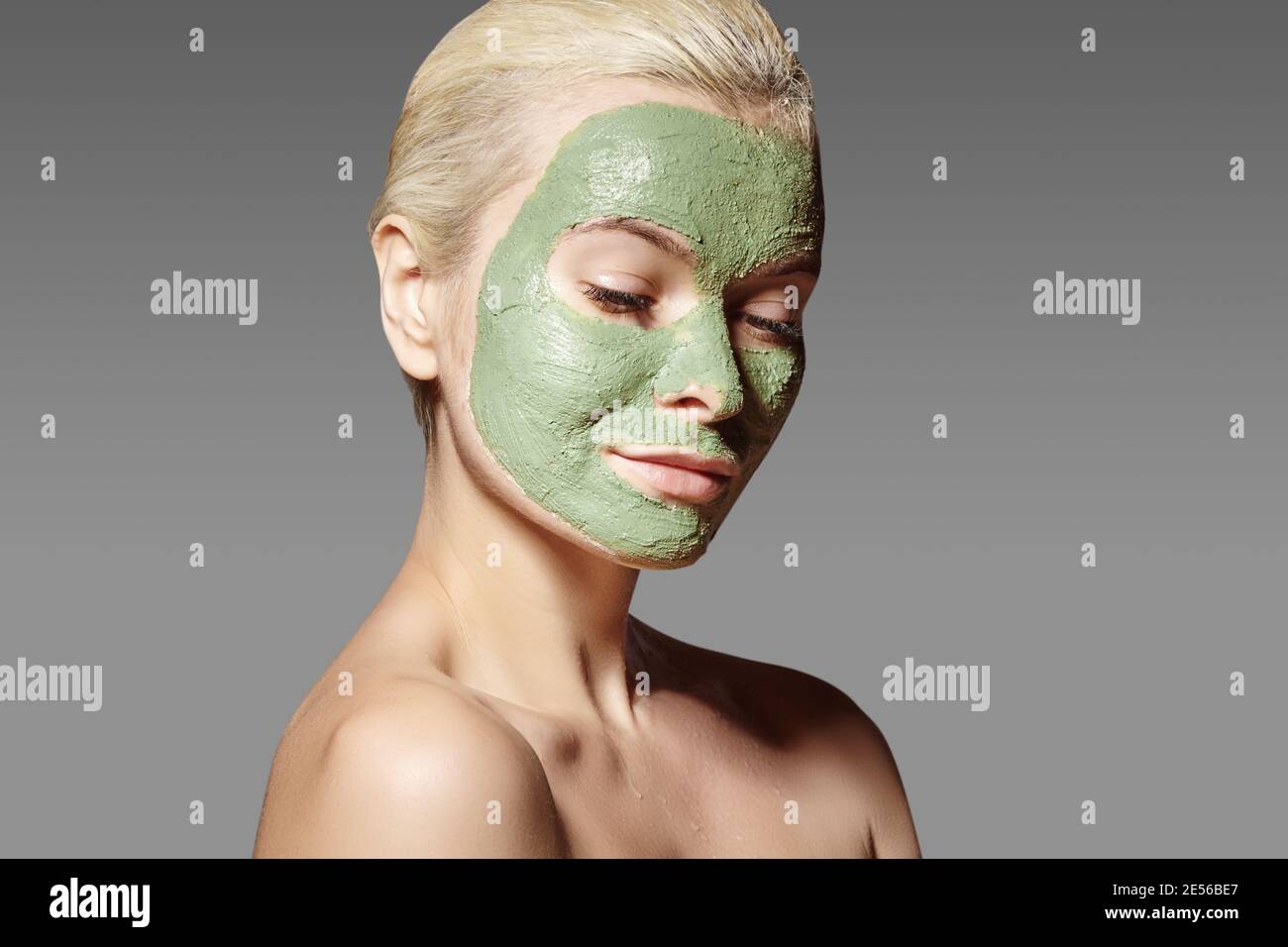 Beautiful Woman Applying Green Facial Mask. Beauty Treatments. Close-up Portrait of Spa Girl Apply Clay Facial mask on grey background. Stock Photo