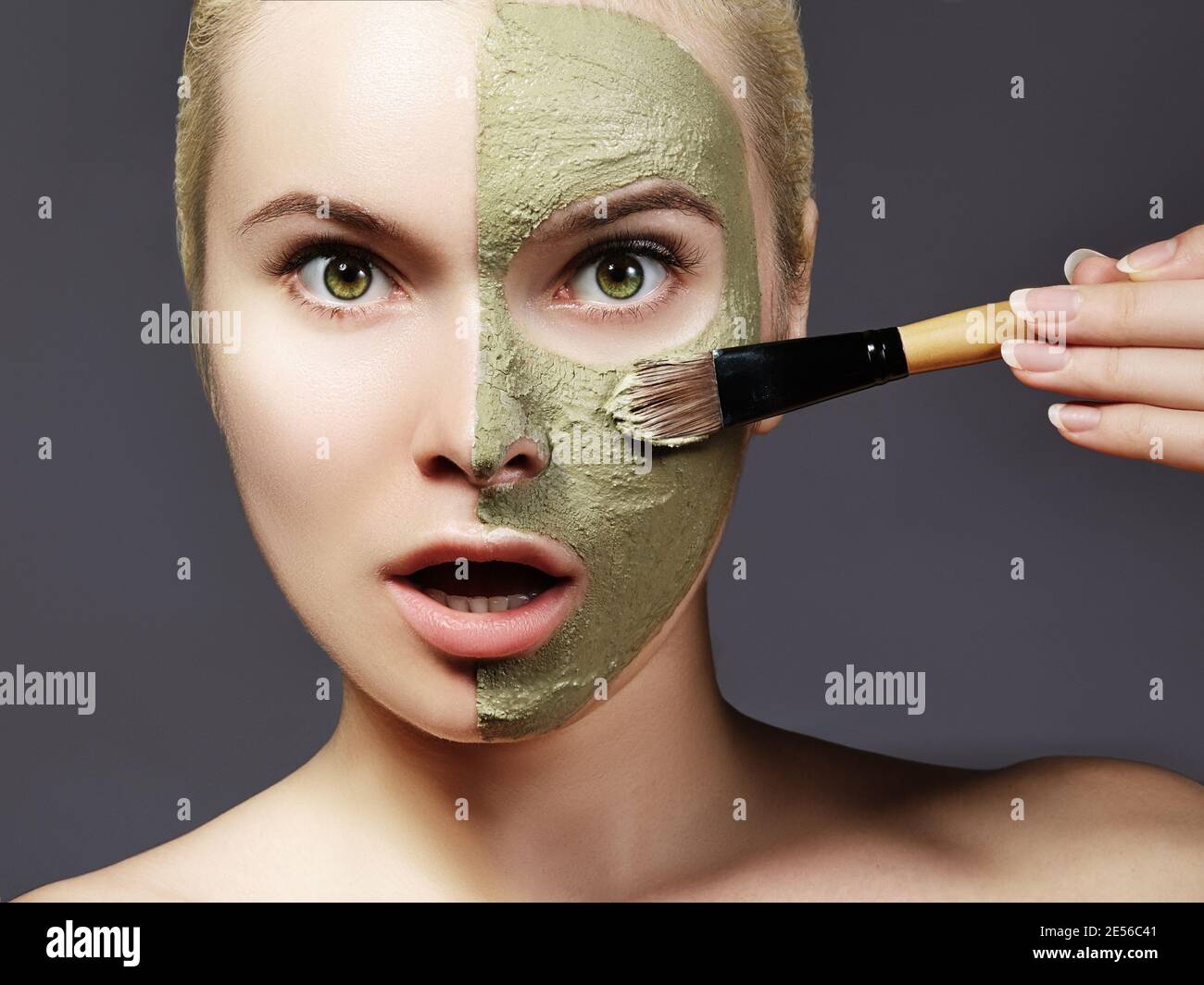 Beautiful Woman Applying Green Facial Mask. Beauty Treatments. Close-up Portrait of Spa Girl Apply Clay Facial mask with brush on grey background Stock Photo