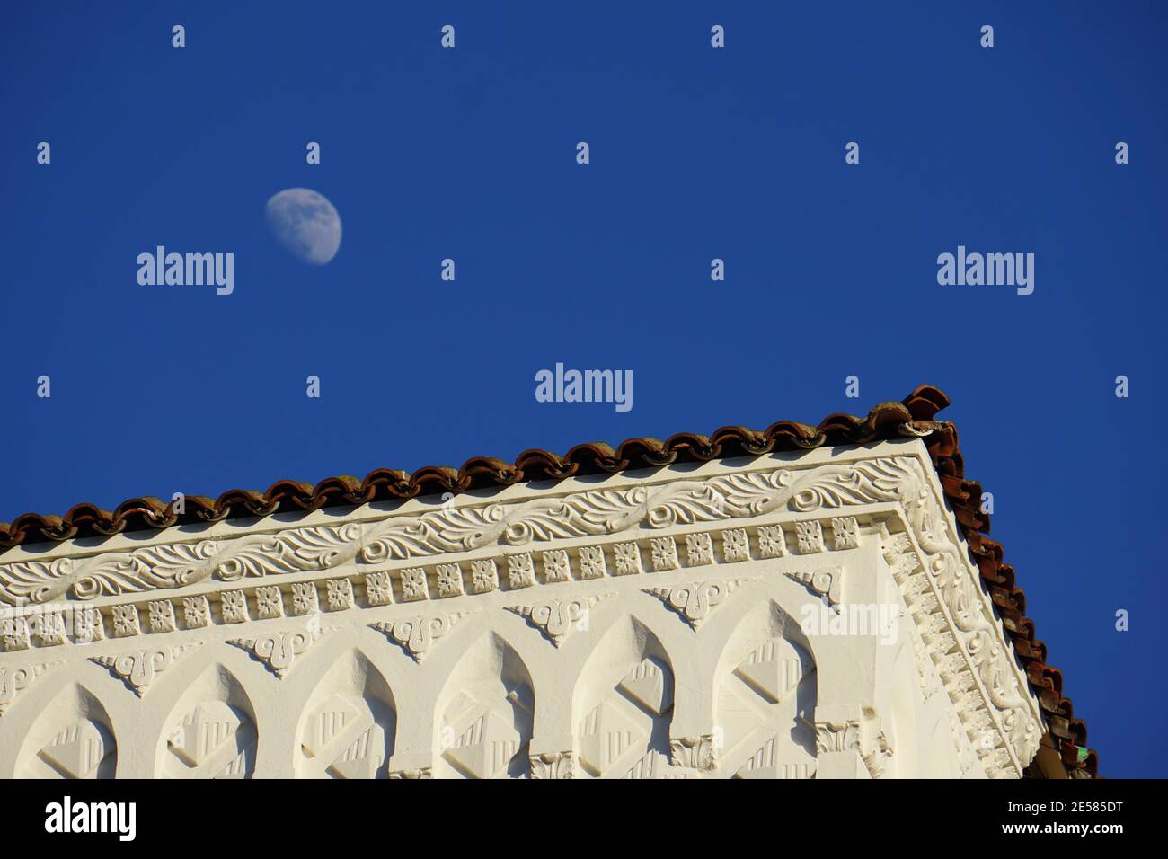 Day moon, white moon, half moon, against a blue sky, hanging above a shingled rooftop. Oakland, California Stock Photo