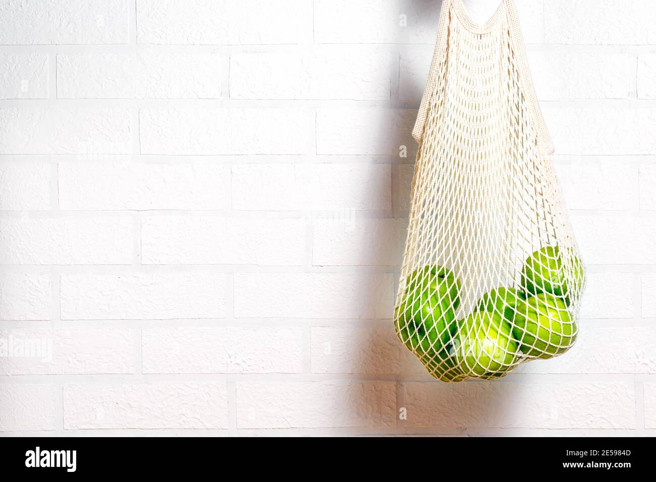 Fresh green apples in eco natural reusable cotton mesh bag. Zero waste concept. White brick background.  Stock Photo
