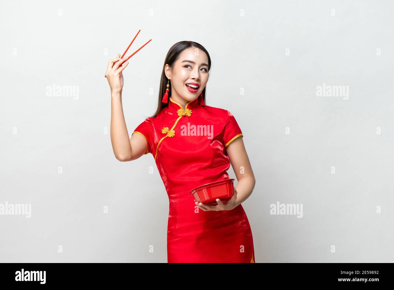 Smiling Asian woman in traditional red Chinese qipao dress holding plastic food box and chopsticks in studio isolated light gray background Stock Photo