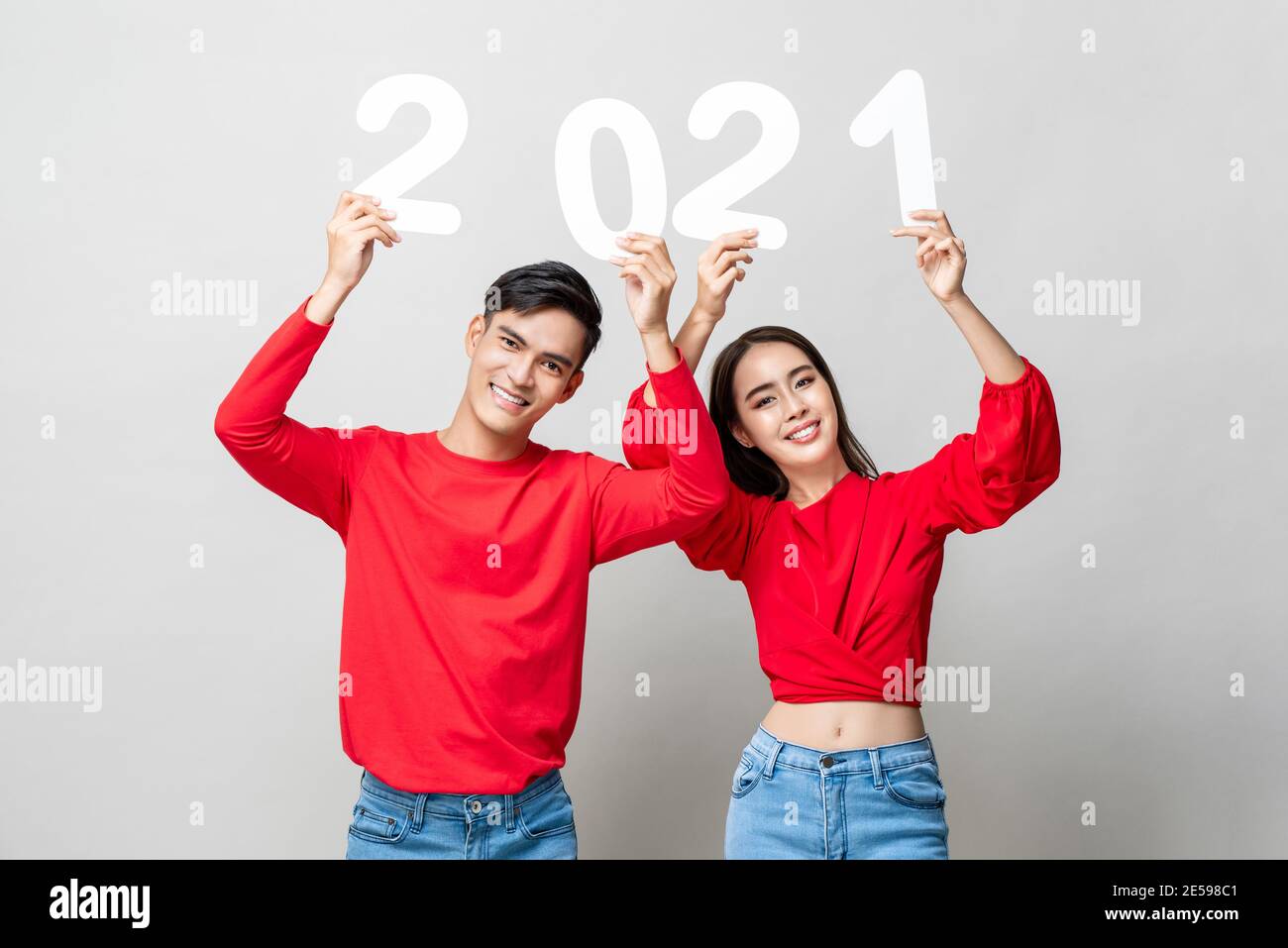 Portrait of smiling happy Asian couple in red casual attire raising 2021 numbers for new year concept on light gray background Stock Photo