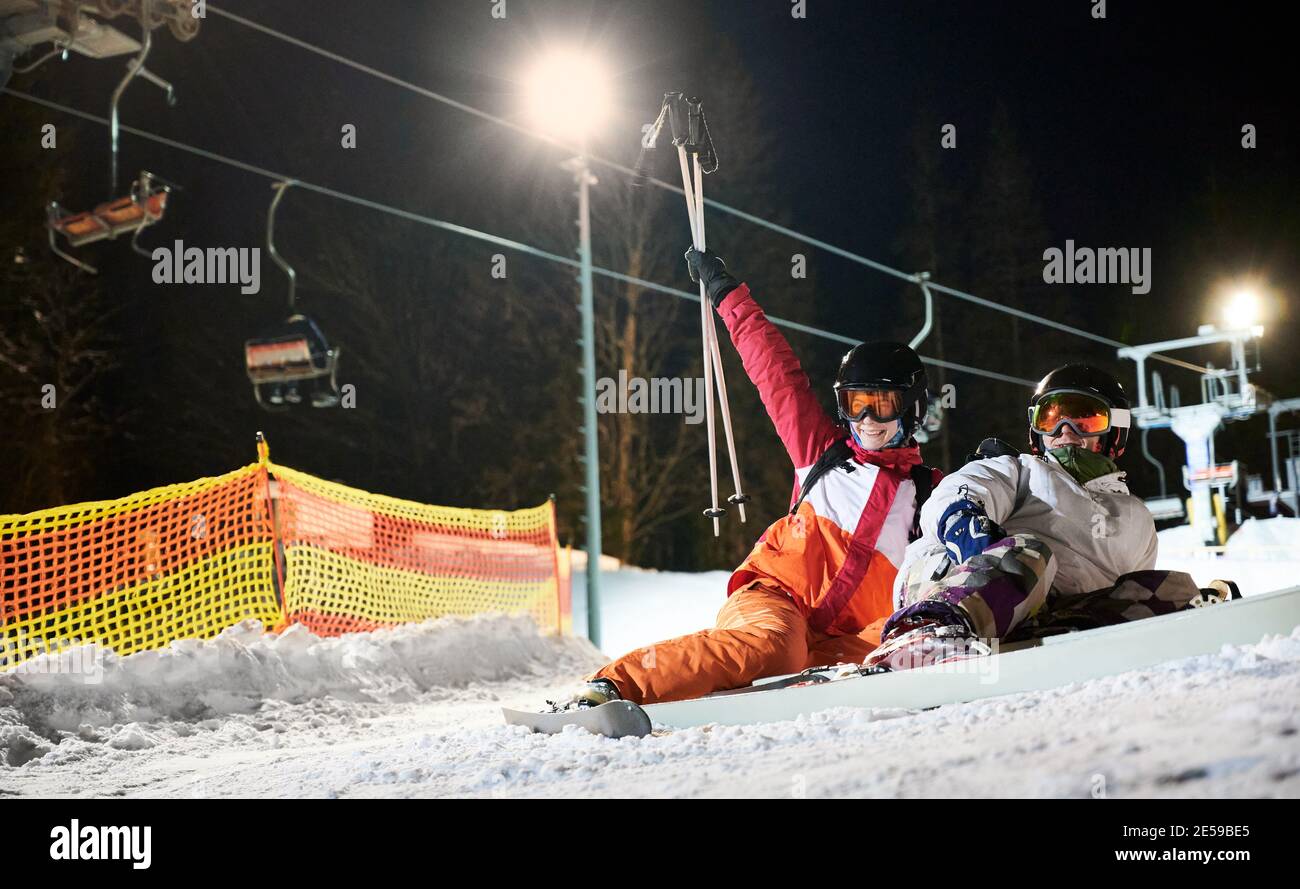 Couple of tourists lying on snowy slope under illuminated chairlift at night, resting after active time spending. Friends at high spirits having fun at ski resort Stock Photo