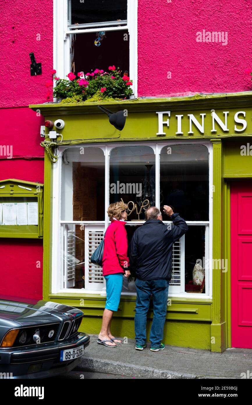 Woman and man look into window of Finns Restaurant, Kinsale Ireland; woman's bright pink jacket matches building walls and window box flowers, contras Stock Photo