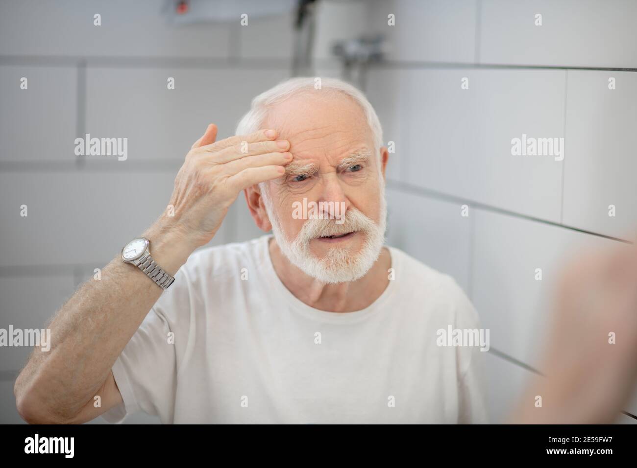 Gray-haired man in white tshirt looking in the mirror and looking worried Stock Photo