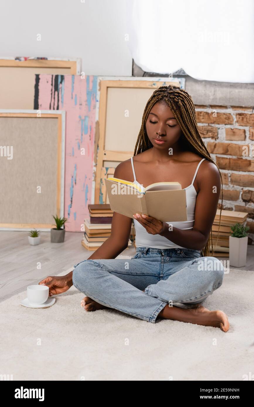 African american woman reading book and holding cup of coffee on carpet ...