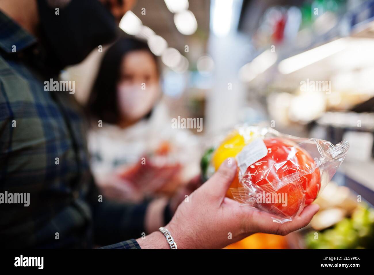 Asian couple wear in protective face mask shopping together in supermarket during pandemic. Taking vegetables from fridge. Stock Photo