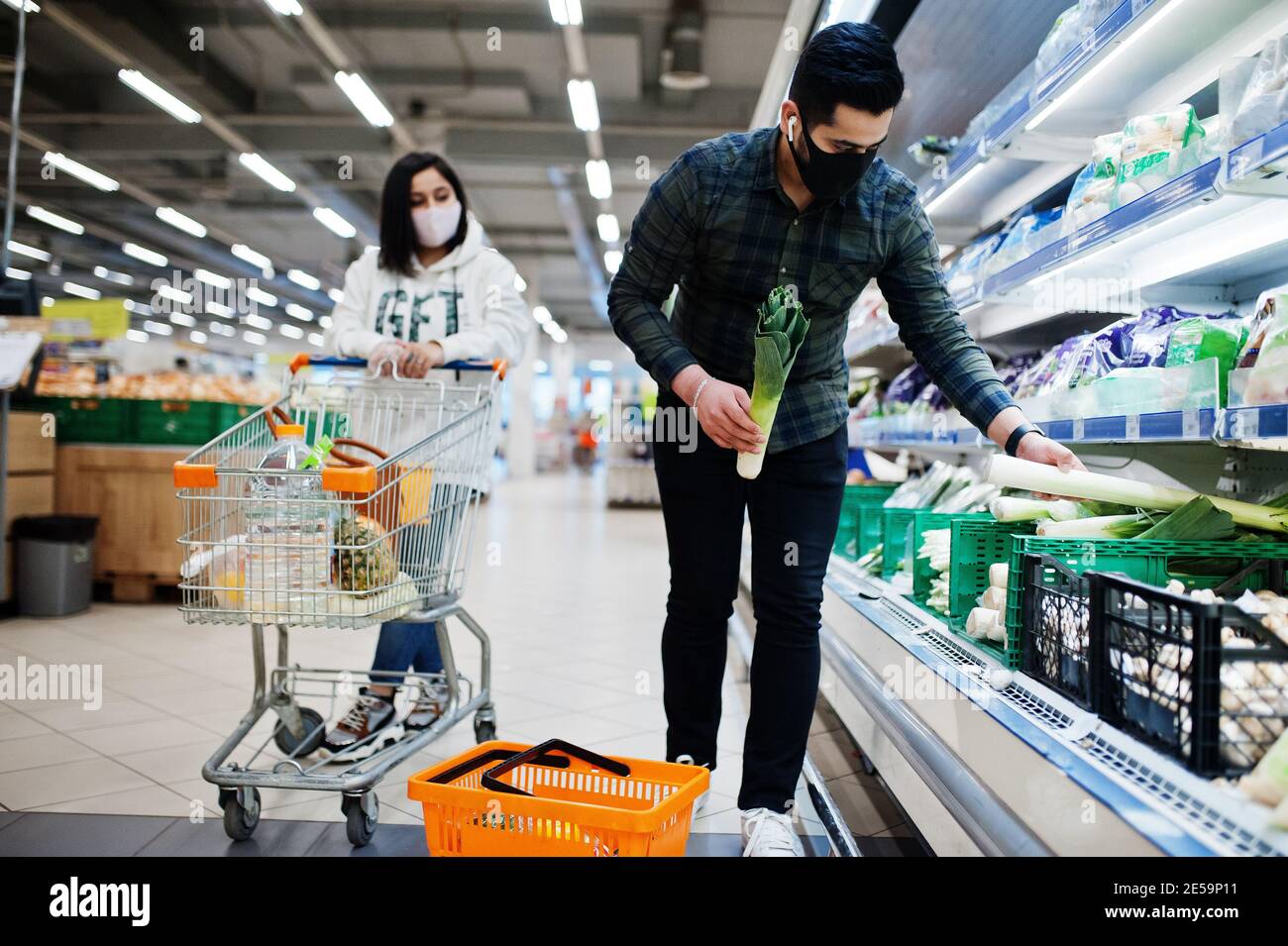 Asian couple wear in protective face mask shopping together in supermarket during pandemic. Taking vegetables from fridge. Stock Photo