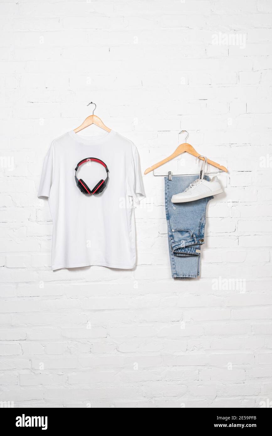t-shirt, wireless headphones, blue jeans and sneaker on hangers near white brick wall Stock Photo