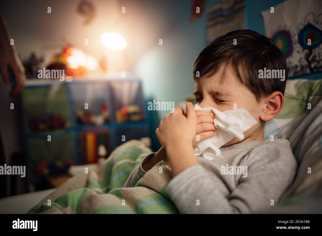 A portrait of a sick child with the flu clearing his stuffy nose. A boy lying in bed blowing his nose. Stock Photo