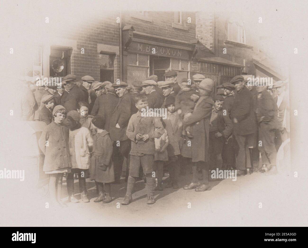 Crowd of men, women & children gathered in the street to listen to a football match being broadcast on the radio at Red Row, Northumberland, c1920/30s. A radio speaker is sticking out of the window of Belle Vue House (on the left of the image). A diagram showing a football pitch divided into numbered squares is on display in the left hand window of M.B. Coxon's shop. The crowd is mostly made up of men in flat caps, with a woman & several girls & boys also in the audience. The first live radio broadcast of a football game took place on 22 Jan 1927 with a match between Arsenal & Sheffield United Stock Photo