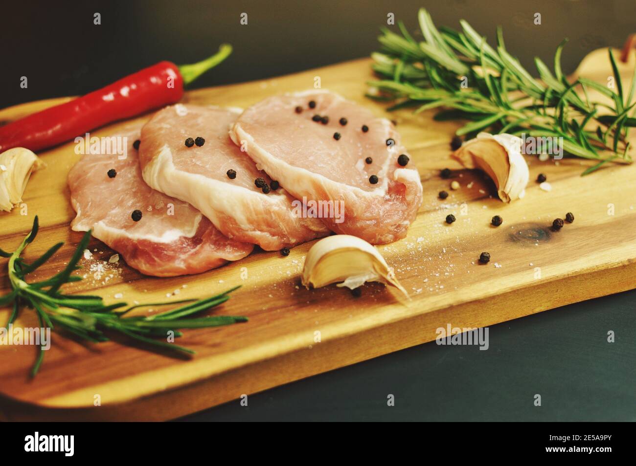 Raw fresh uncooked sliced pork meat fillet dish with rosemary, pepper, salt, red chili pepper, garlic on wooden board and black background. Stock Photo