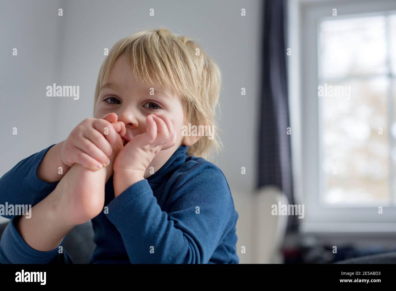 Cute blond toddler child, sucking his foot thumb, making funny faces, laughing Stock Photo