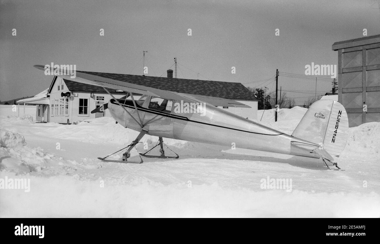 Vintage 1950s American black and white photograph of a Cessna 120 light aircraft, serial number NC2842N. Aircraft is fitted with skis and is parked on a snow covered area alongside some buildings. Stock Photo