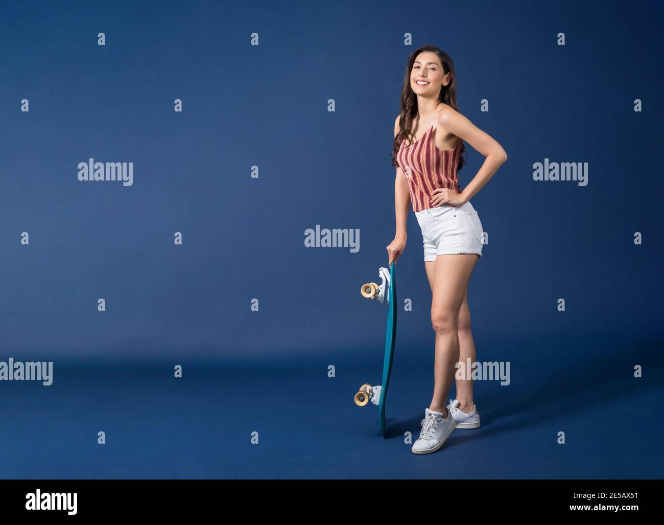 Happy young Asian woman standing and holding surfskate or skateboard on blue color background, exercise and sport, trendy and extream sports concept Stock Photo
