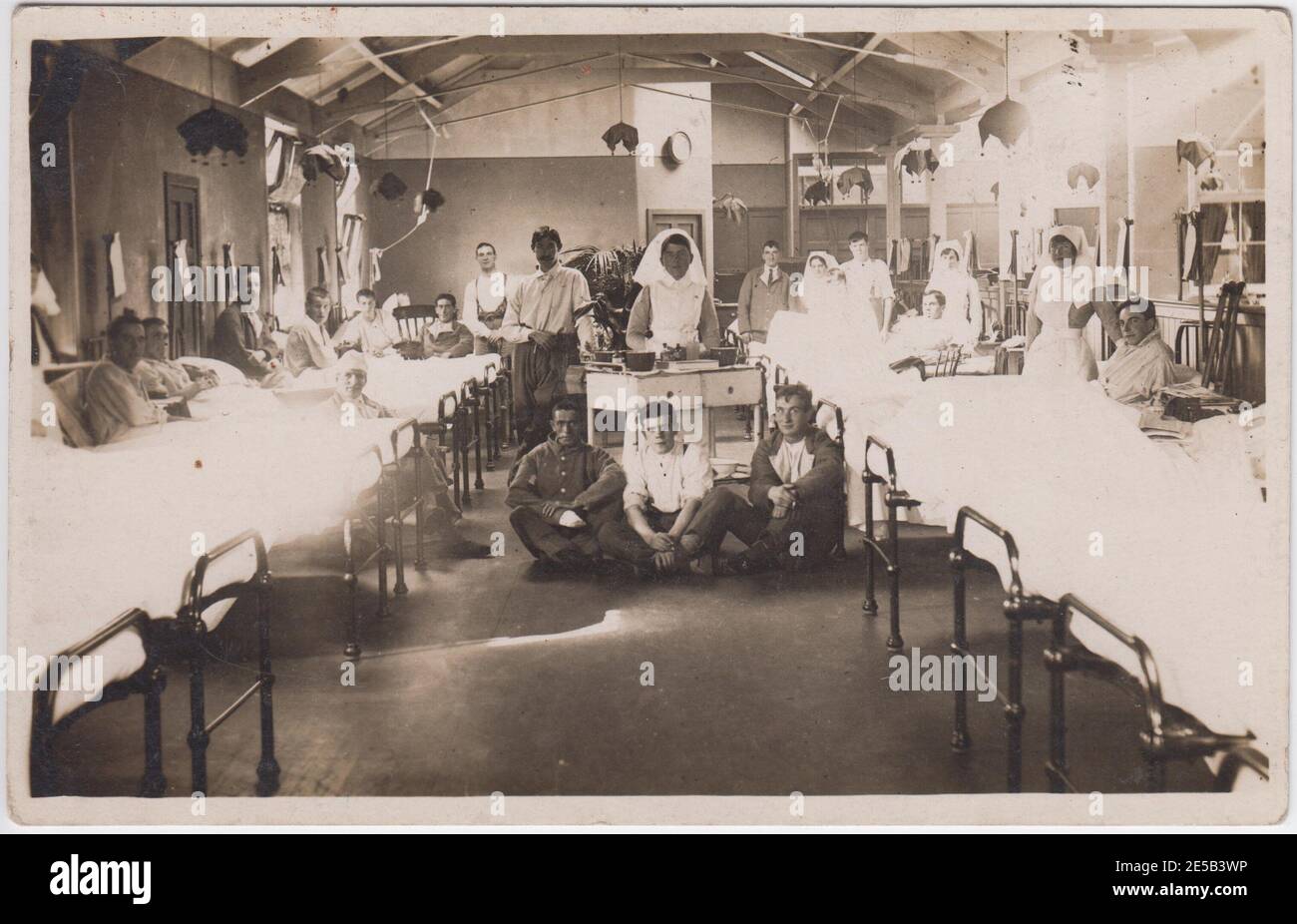 Ward of a First World War military hospital. Patients, many wearing hospital blue uniforms, are sitting up in their beds, sitting in the central aisle and standing towards the back of the room. Nurses are standing by the beds and behind a trolley in the centre aisle. Stock Photo