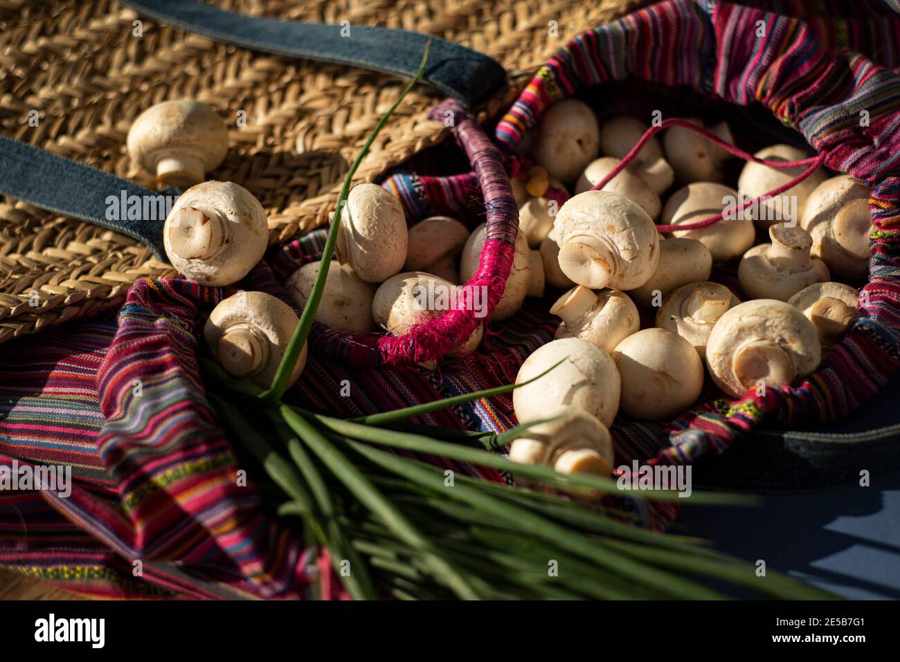 Champignons and spings onions are poured out of a wicker boho bag. The theme of healthy eating and sustainable living. Stock Photo