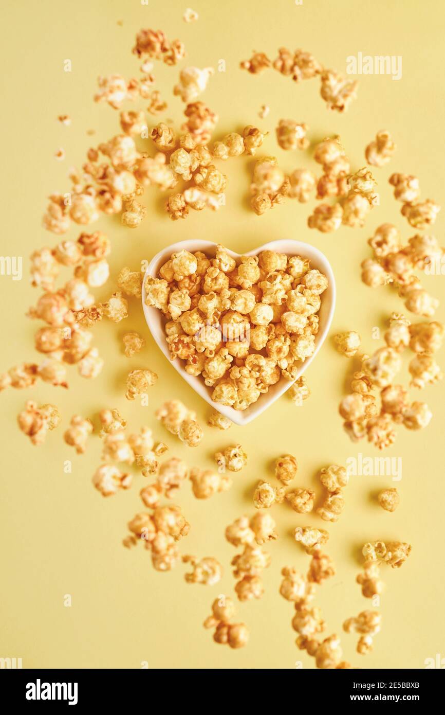 Flying delicious sweet popcorn with caramel in white ceramic heart plate, isolated on trend color yellow background. Stock Photo