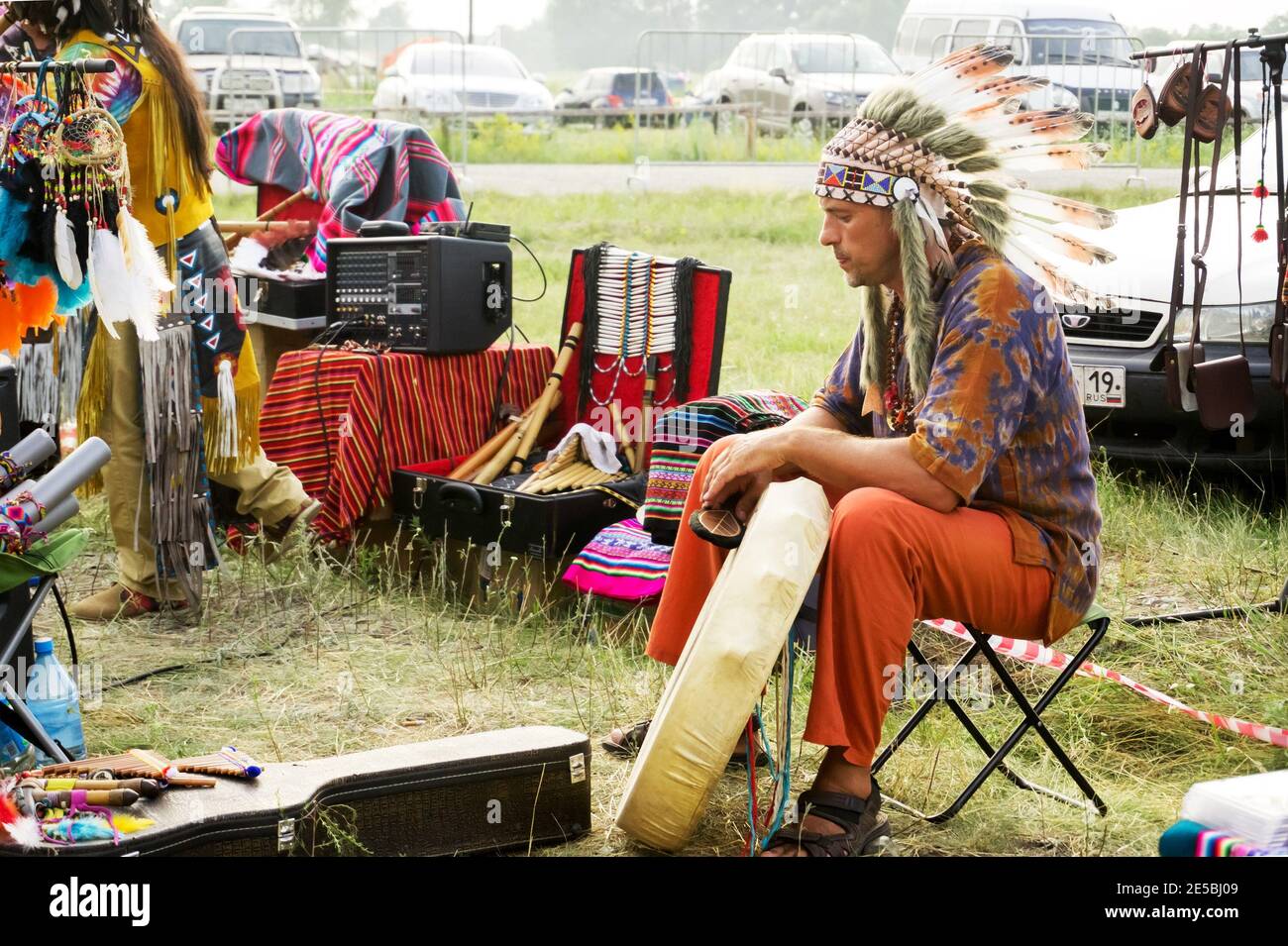 A man in a headdress with feathers and a tambourine sits near a car during the annual Intl festival of music and crafts World of Siberia. Stock Photo