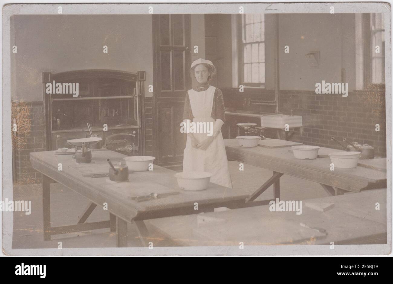 Edwardian servant standing in a large kitchen. Bowls and pans are on the tables around her. A sink and range are in the background. Stock Photo