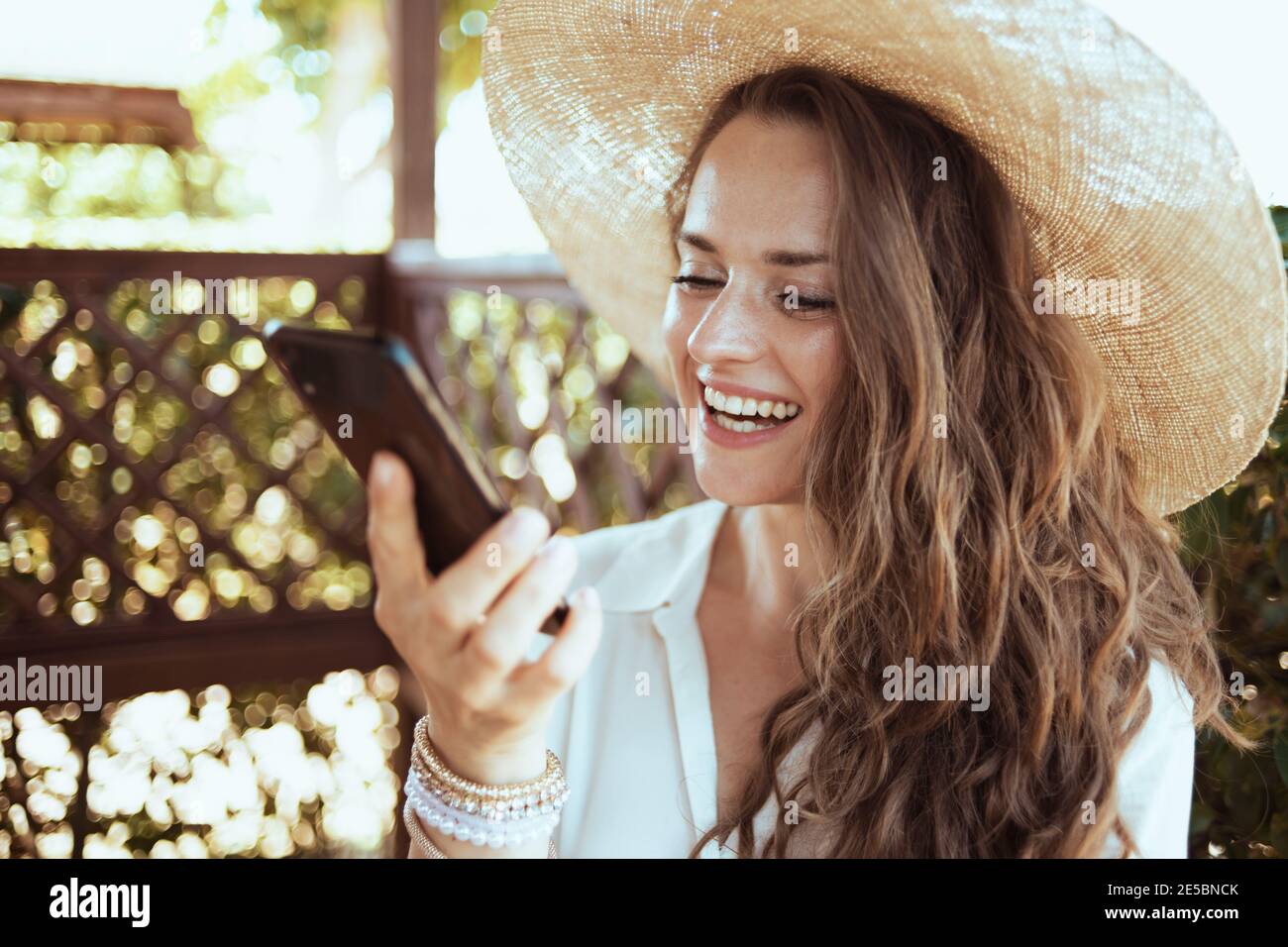 happy elegant middle aged woman in white shirt with hat having video call on a smartphone in the terrace of guest house hotel. Stock Photo