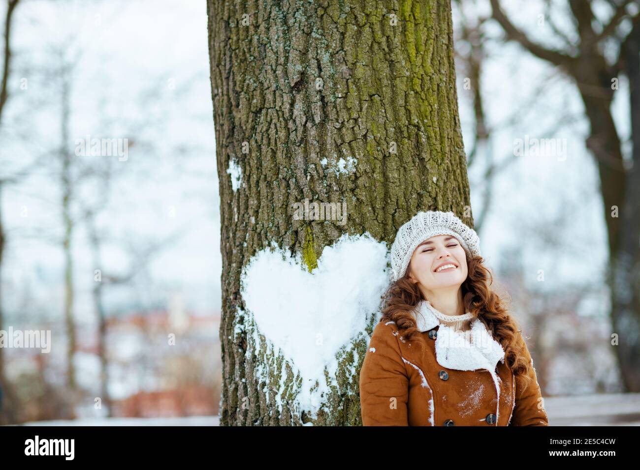 happy elegant female in a knitted hat and sheepskin coat near tree with snowy heart outdoors in the city park in winter. Stock Photo